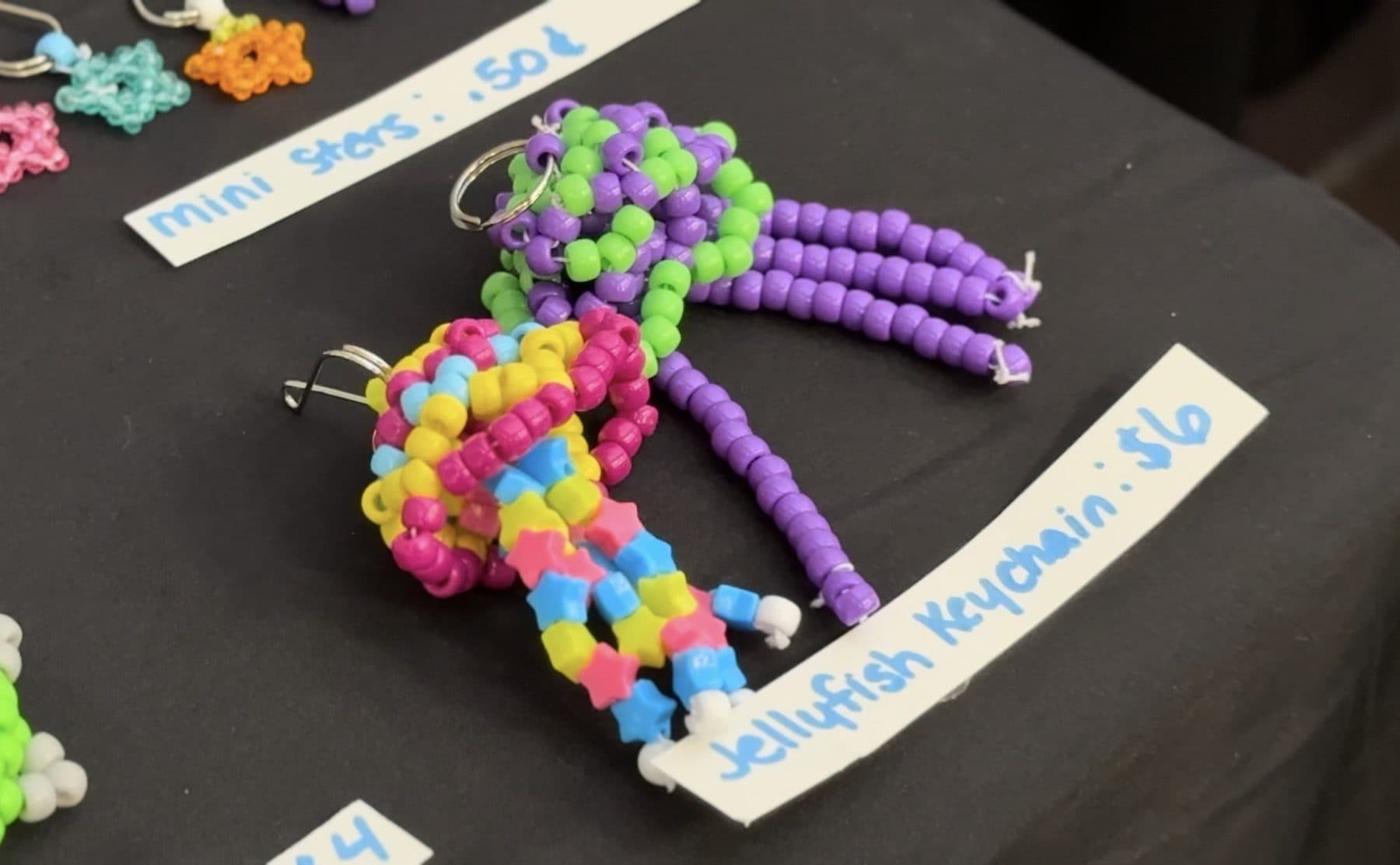 Beaded jellyfish keychains on a stall table beside hand-lettered price labels reading mini stars 50 cents and jellyfish keychain $6.