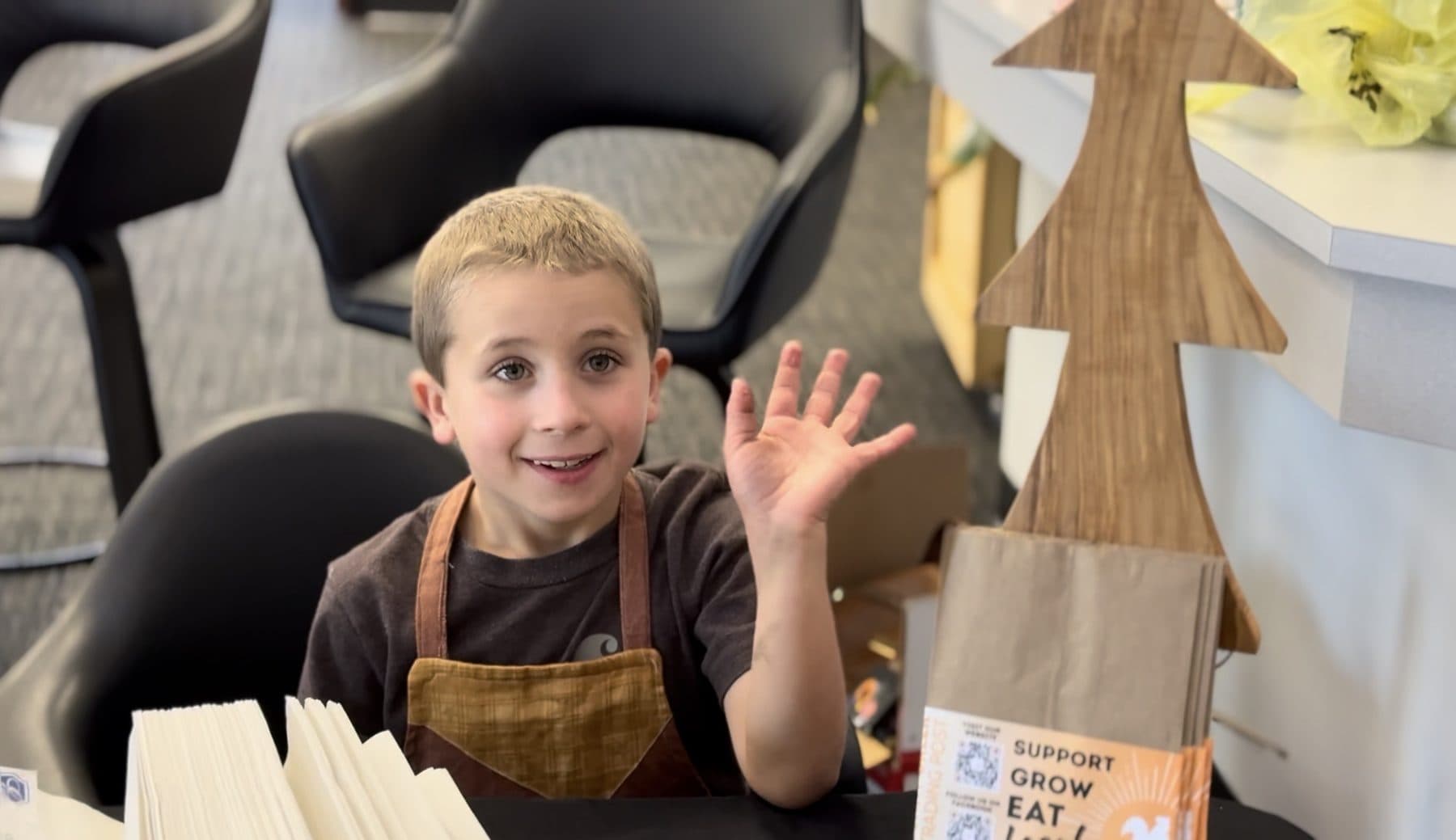 A young boy in a work apron waving from behind his table of handmade goods.